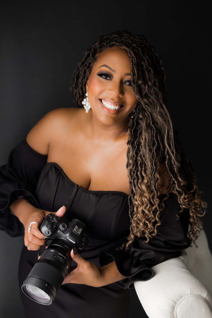 A photo of Michelle Harris, a black woman with long braids over her shoulder and wearing a black strapless dress. She is smiling at the camera, and is holding a camera in her hands.