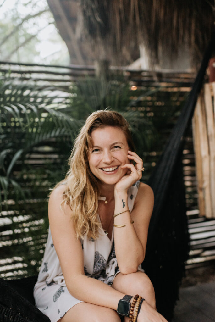 Sara, a white woman with blonde hair, leans her chin on her hand and props her elbow on her knee. She smiles at the camera and is sitting in front of a palm frond wall.