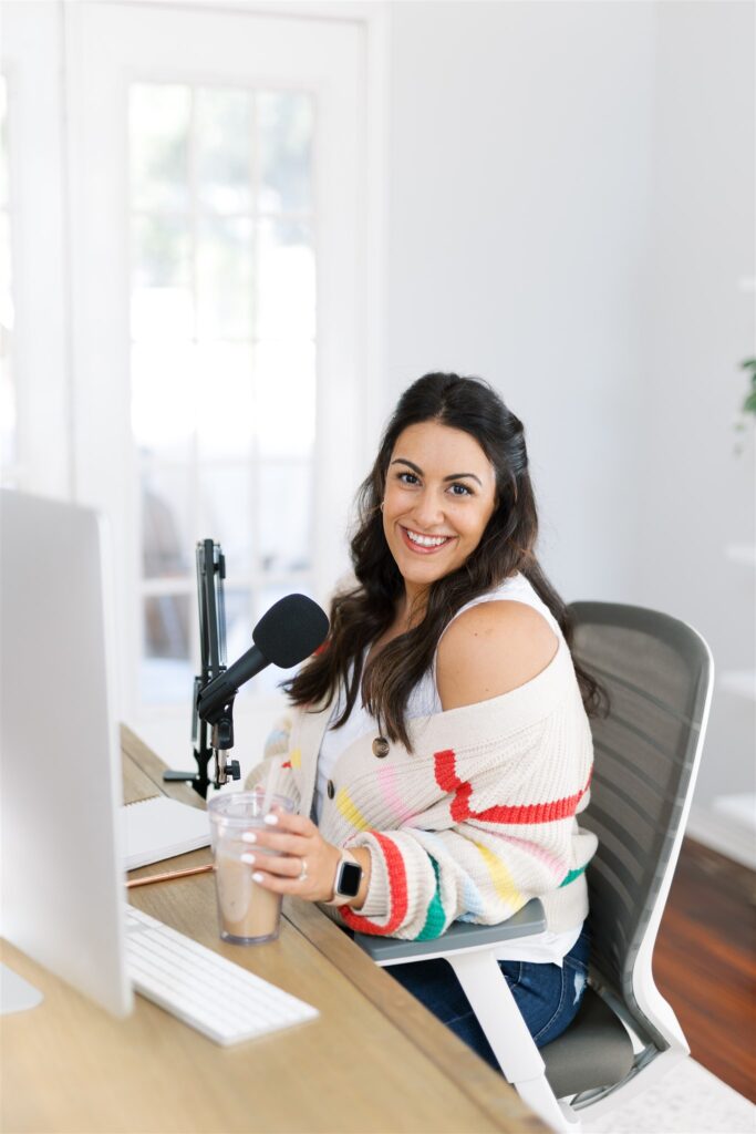 Laylee Emadi holds an iced coffee and sits and a computer desk in front of a podcast microphone.