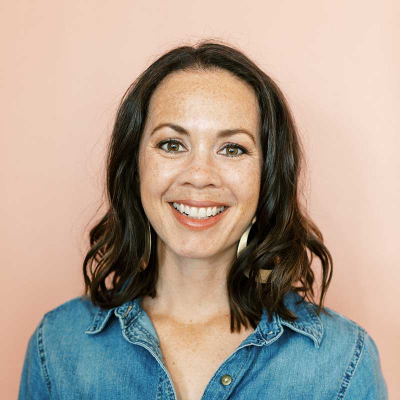 A smiling woman with shoulder-length dark hair wearing a denim shirt, photographed against a soft peach background; author photo for guest contributor Tayler from Enji.