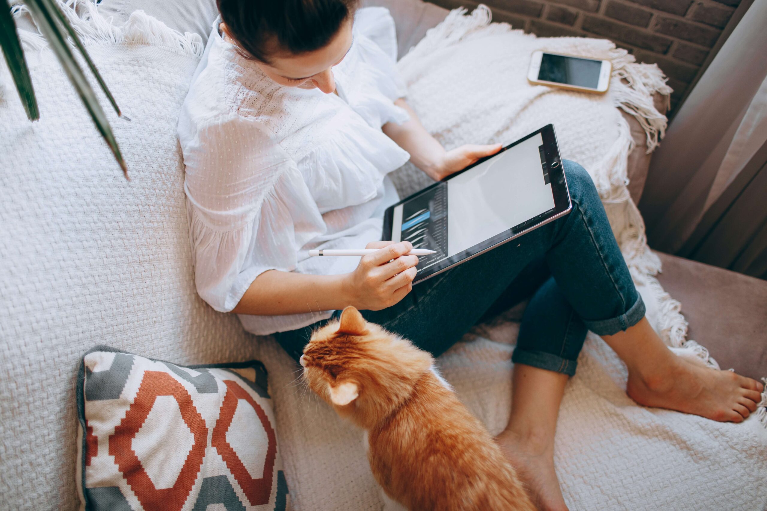 A woman sitting on a couch using a tablet and stylus to work on a project, with a ginger cat beside her; cozy home workspace representing content creation and marketing tasks.