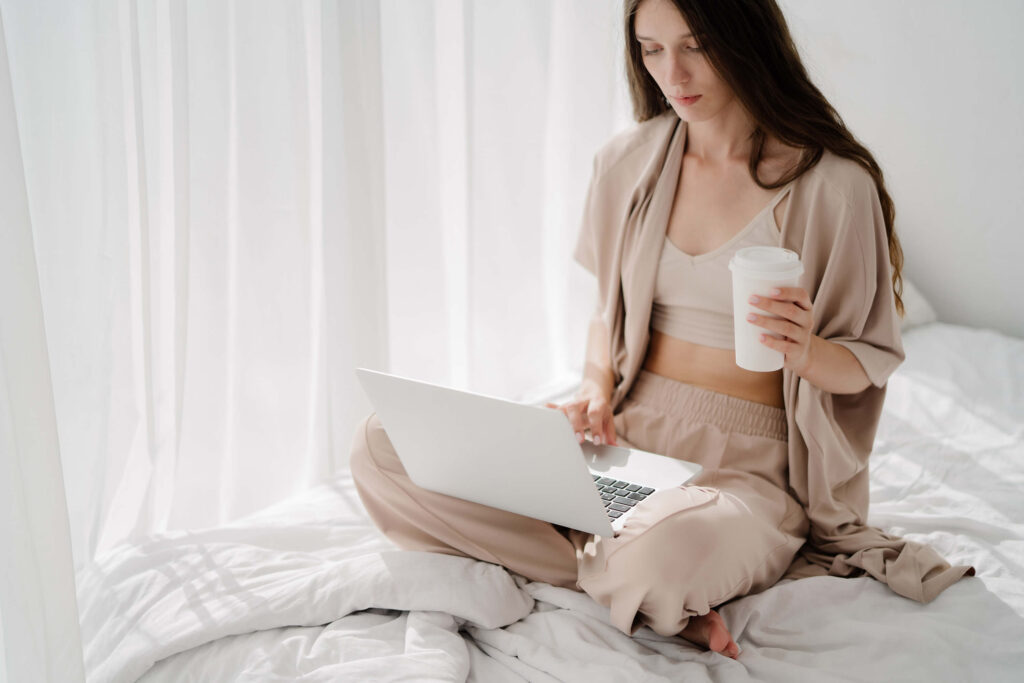 A woman sitting on a white bed with a laptop and coffee, representing a wedding professional working on Flodesk email marketing in a relaxed setting.