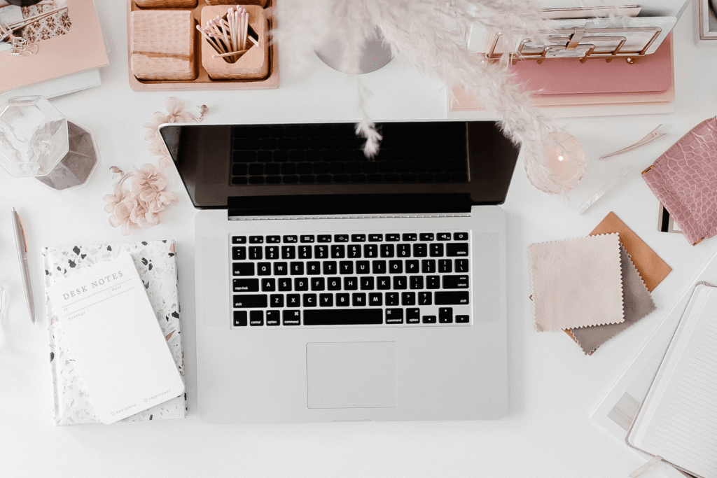 An overhead view of an open laptop on a white desk surrounded by pink and gold stationery, pampas grass, fabric swatches, and a terrazzo notebook — tools for building a more intentional client experience as a wedding professional.