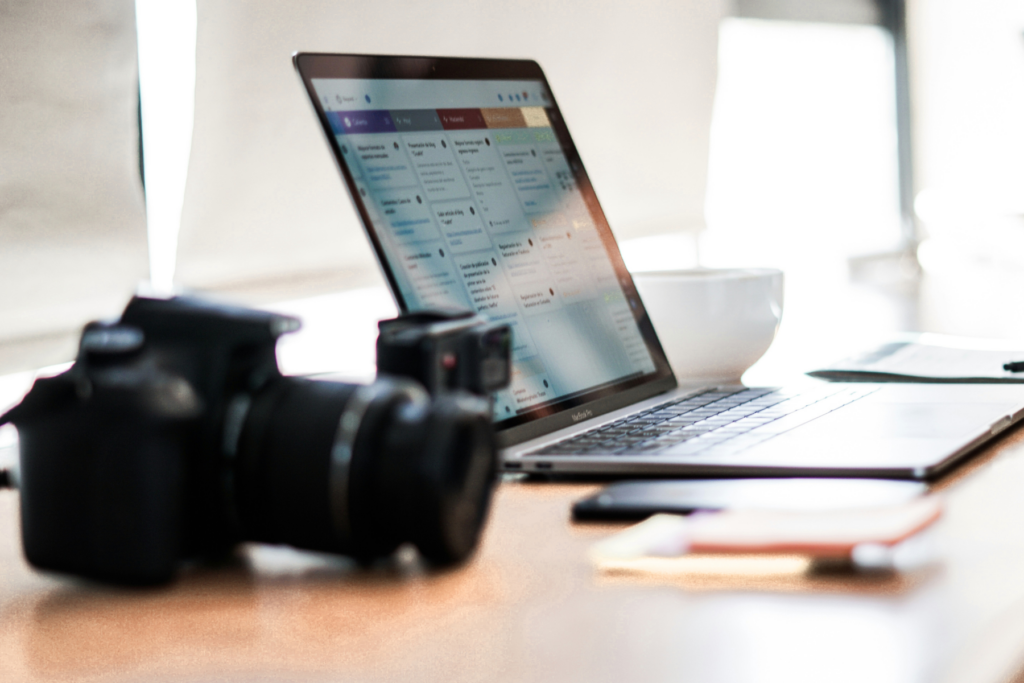 A DSLR camera sitting in front of an open laptop displaying a project management board on a wooden desk — a wedding photographer planning their wedding day timeline and workflow before the season starts.