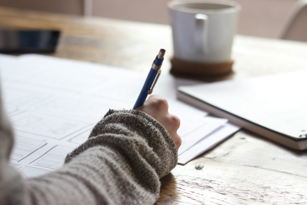 A close-up of a hand in a grey knit sleeve writing on printed documents at a wooden table with a notebook and coffee mug nearby — a wedding photographer reviewing and annotating a wedding day timeline before the event.