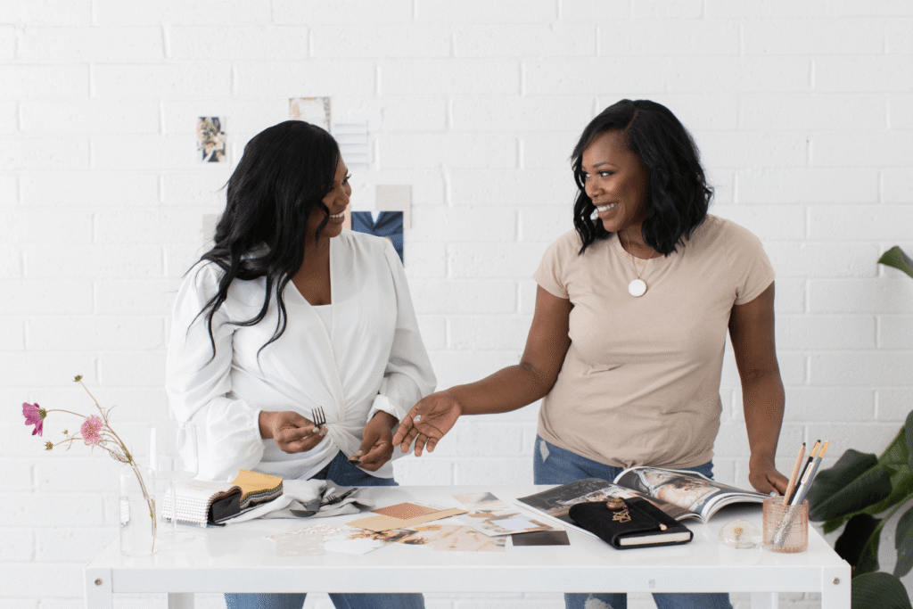 Two Black women smiling and talking across a white desk covered with fabric swatches and photos — representing the confident, prepared client meeting that sending a wedding photographer welcome packet makes possible.