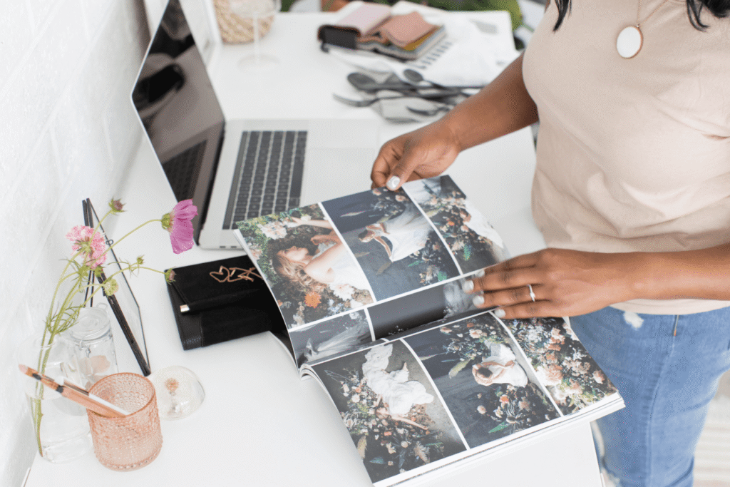 A person flipping through a page of wedding photos in a printed welcome guide at a white desk with a laptop nearby — showing how a well-designed wedding photographer welcome packet can include visual examples that set expectations before the wedding day.