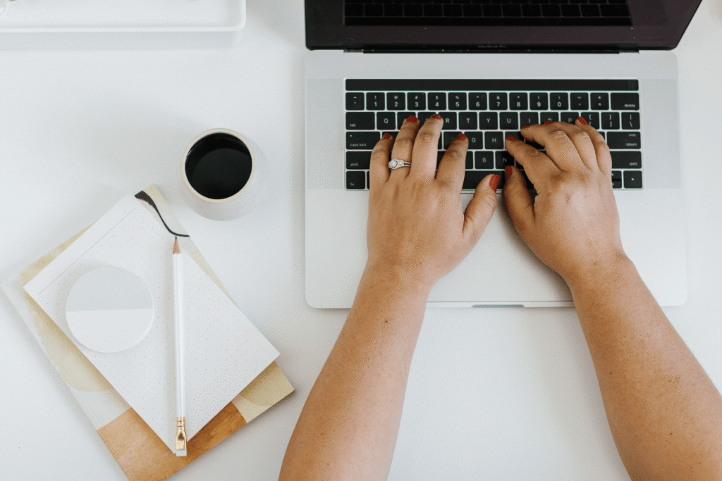 An overhead view of two hands with red nails and a diamond ring typing on a MacBook laptop, with a black coffee cup, a pencil, and a dotted notebook on a white desk — building out a wedding photography client questionnaire.