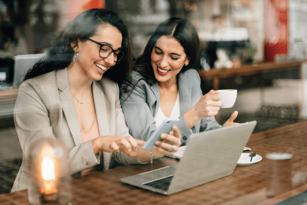Two women smiling and looking at a smartphone together at a café table with a laptop and coffee — the kind of relaxed, engaged conversation that good wedding photography consultation questions make possible.