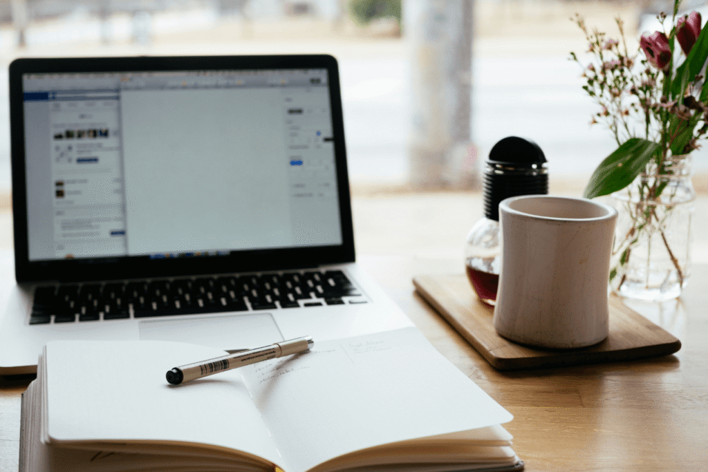 An open notebook with a pen resting on it beside an open laptop, a white ceramic mug, and a small vase of pink flowers on a wooden table — a quiet workspace for preparing wedding photography consultation questions.