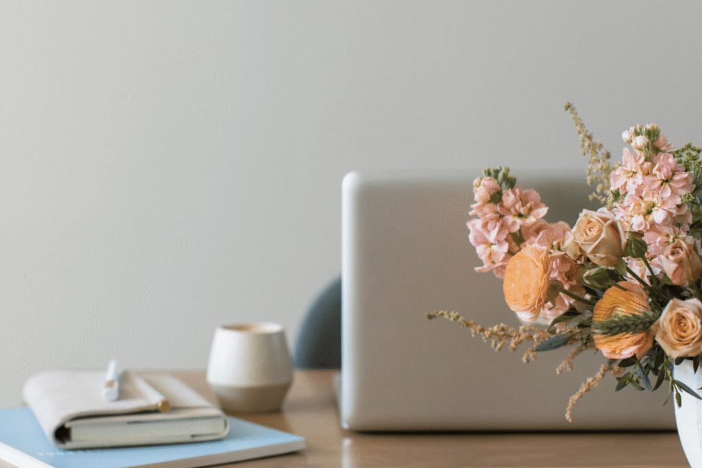 A wooden desk with an open laptop, a small ceramic cup, a notebook with a pen, and a vase of pink and peach flowers — the kind of calm workspace where a wedding photography workflow actually gets built.