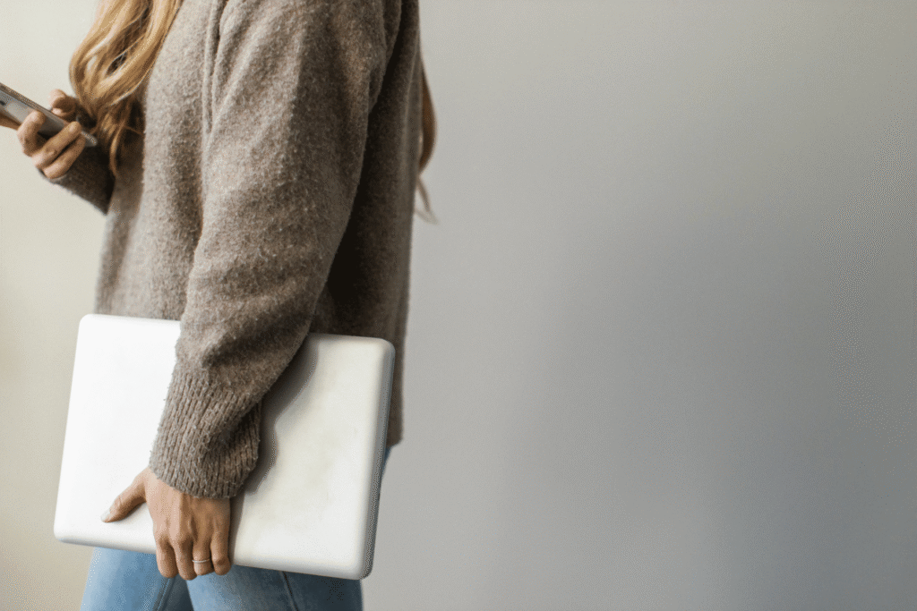 A person with blonde hair wearing a brown knit sweater holding a closed laptop under one arm and a smartphone in their other hand, managing their wedding photography workflow on the go.
