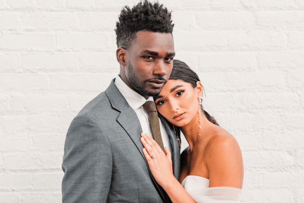 A couple in wedding attire standing close together against a white brick wall, the bride leaning her head on the groom's chest with her hand on his lapel — the kind of intimate portrait that helps wedding photographers get more referrals through word of mouth.