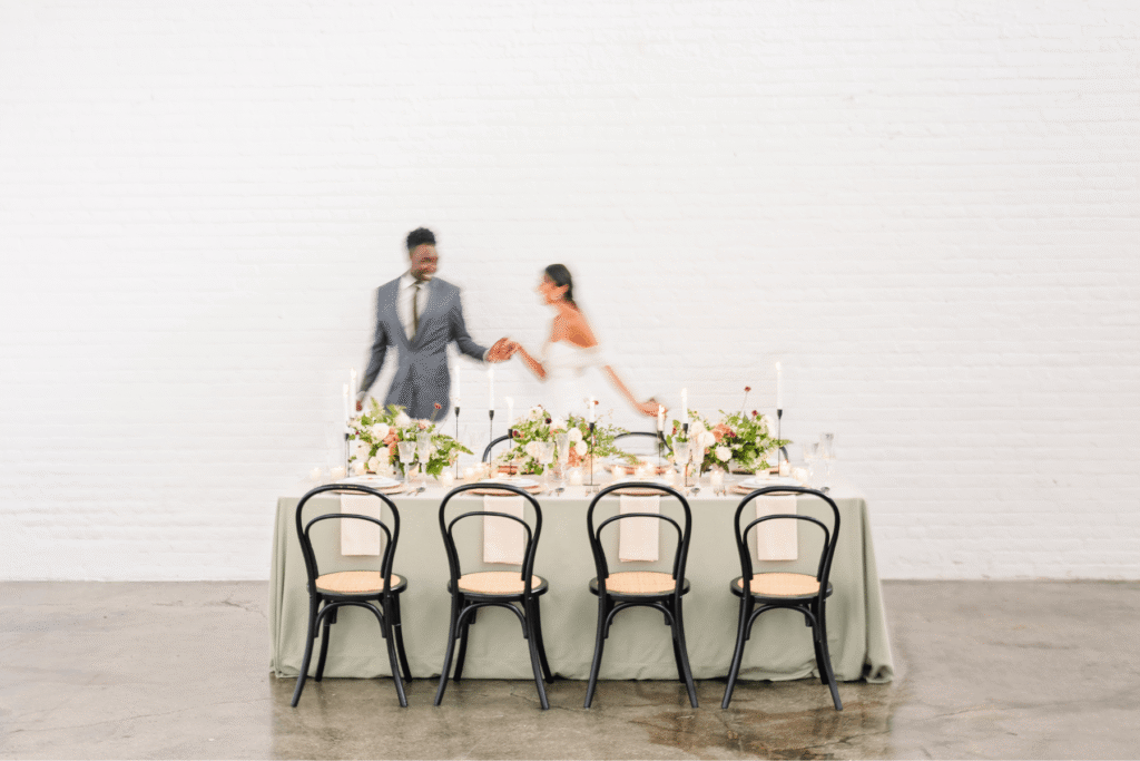 A motion-blurred couple in wedding attire holding hands and walking behind a styled reception table with floral arrangements, candles, and black bentwood chairs against a white brick wall — a memorable wedding moment that leads to referrals for the photographer who captured it.