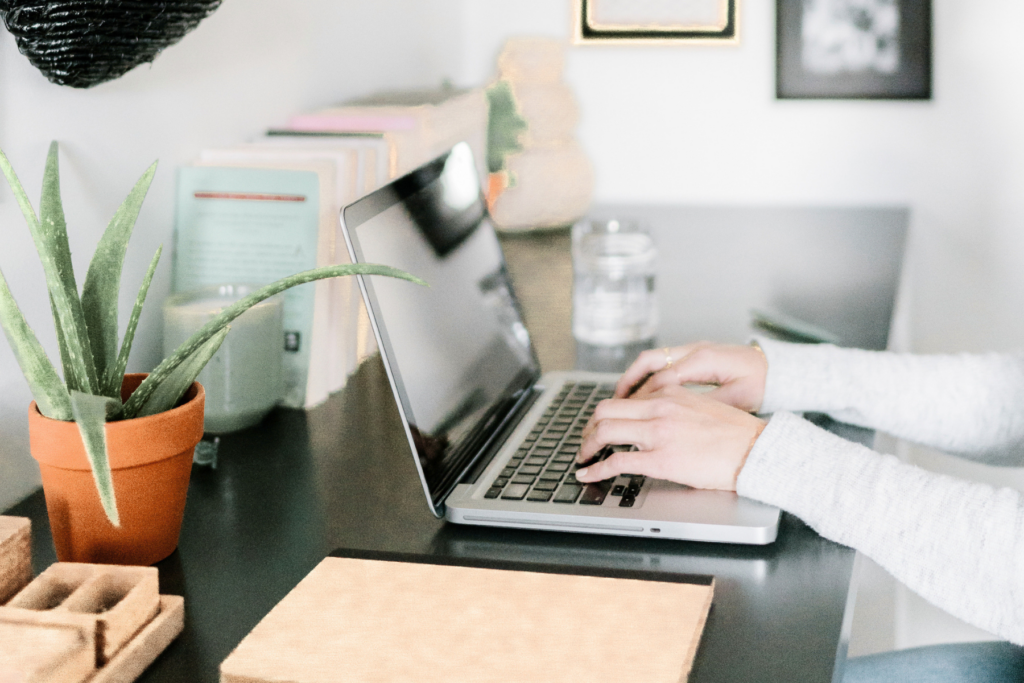 A person in a white sweater typing on a laptop at a dark desk with an aloe plant, books, and a notebook nearby — a wedding photographer building out their wedding day weather backup plan before peak season.