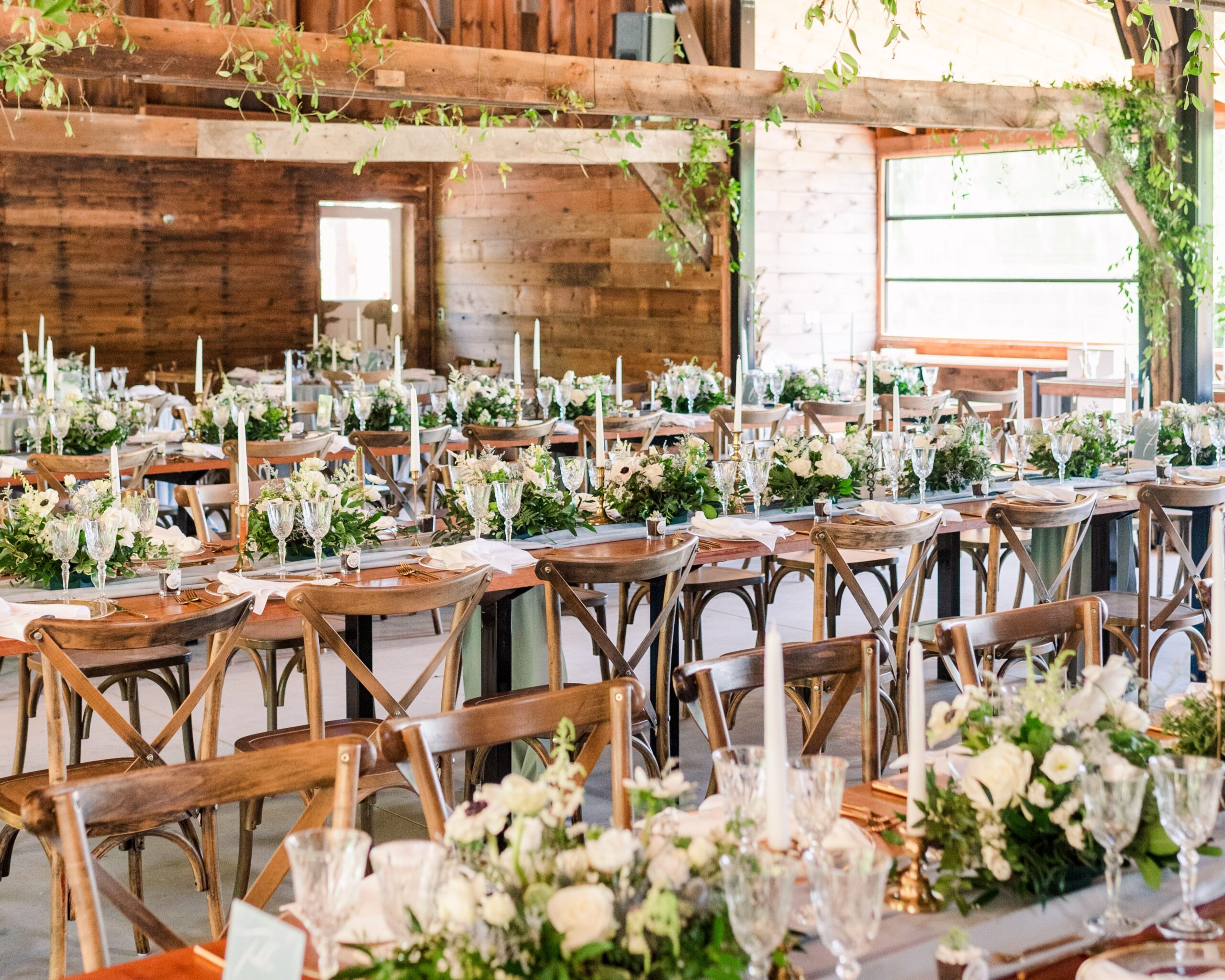 A wedding reception setup inside a barn. The wood tables are filled with greenery and white flowers, white candles, and blue drapery.