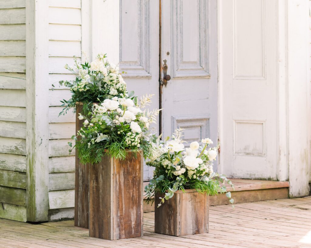 A wedding reception setup inside a barn. The wood tables are filled with greenery and white flowers, white candles, and blue drapery.