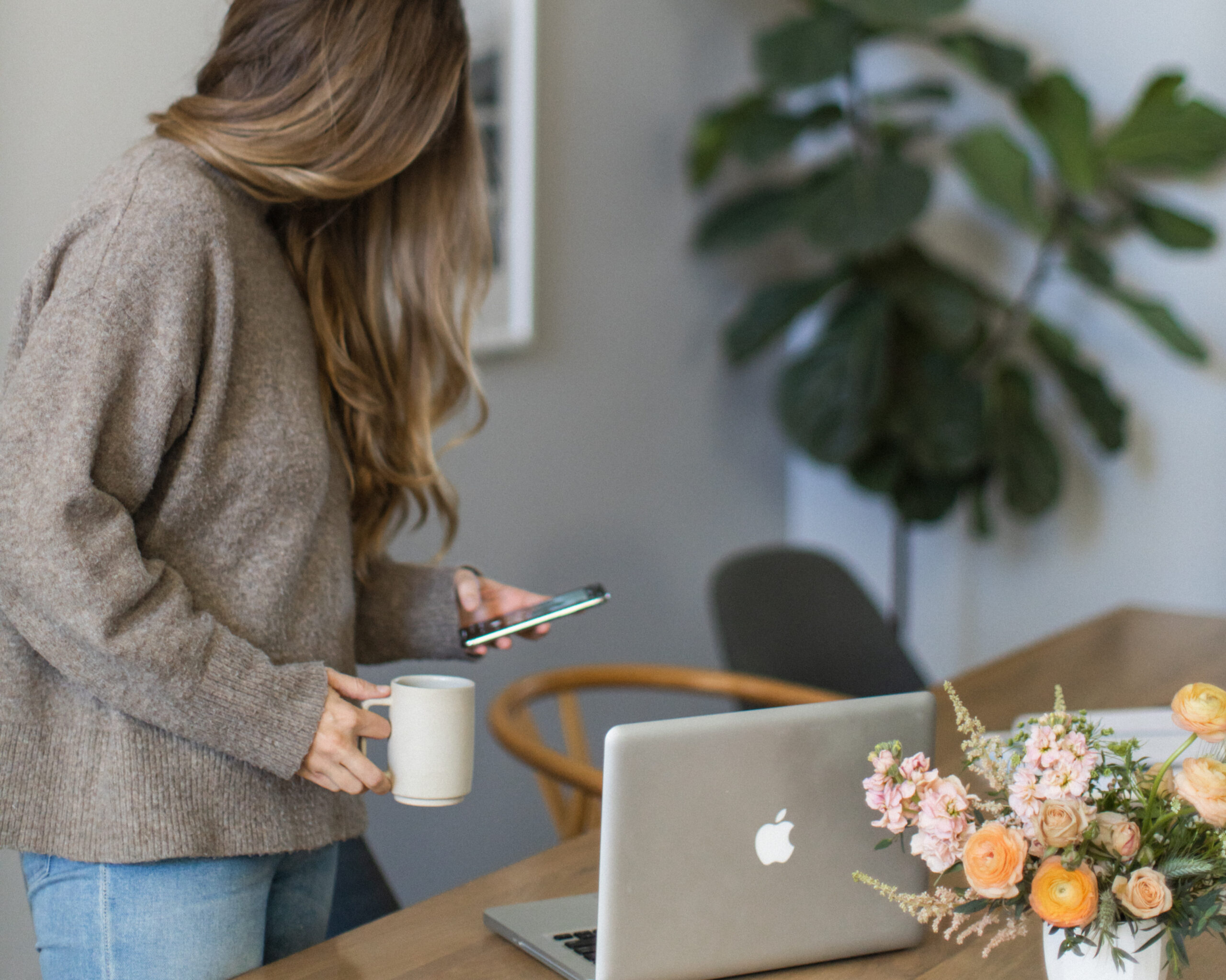A woman holds a coffee mug in her hand and scrolls on her phone with the other hand. She's a fully booked photographer walking towards her laptop at the table.