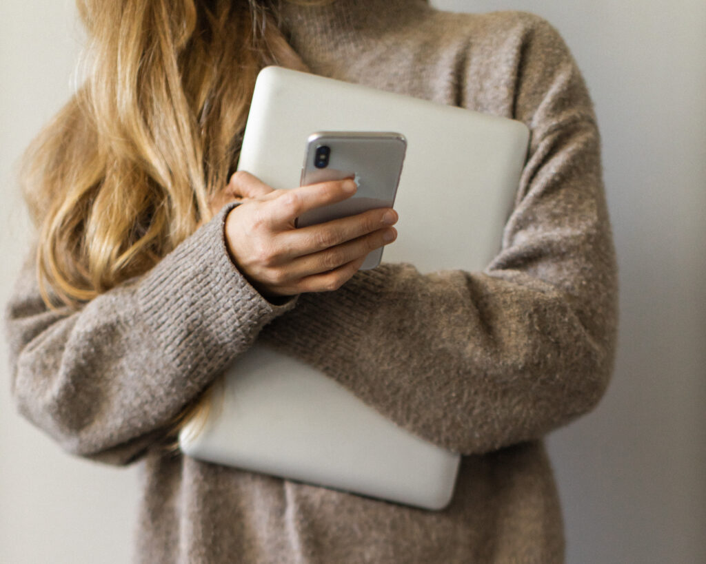 A woman with long blonde hair holds a laptop against her chest with one hand and holds a phone in the other hand as she scrolls on it.