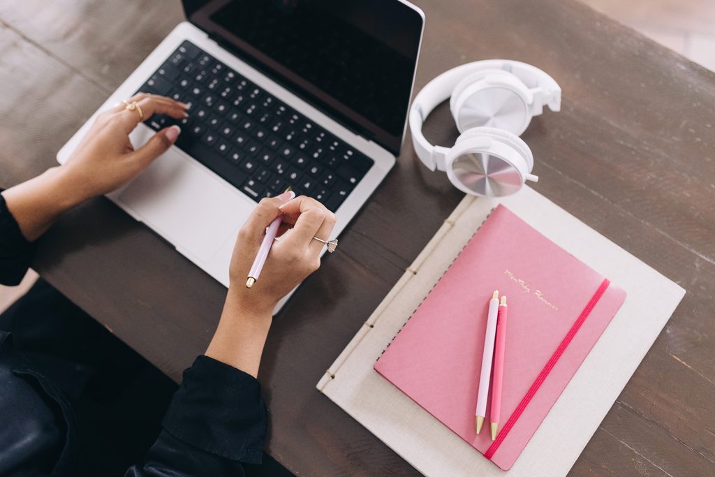 A woman's hands hover over her laptop. She has a notebook and pen beside her.