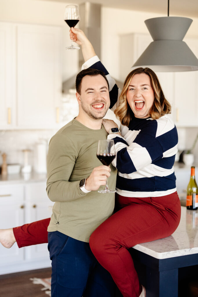 Tia sits on a counter and Cameron stands in front of her. They're both holding glasses of wine and smiling at the camera. Photo is used in Cameron and Tia's bio.