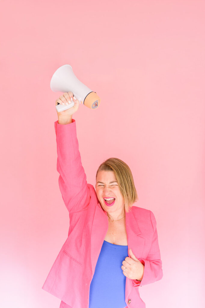 Erin from Mabely Q, a white woman with short blonde hair, is holding a megaphone and throwing her hand in the air. She has a big smile on her face and is standing in front of a pink background.