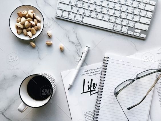 a computer keyboard, a mug of coffee, a notebook, glasses, a pen, and a dish of nuts, are all on a marble table. used in a blog post about consistency in your photography business