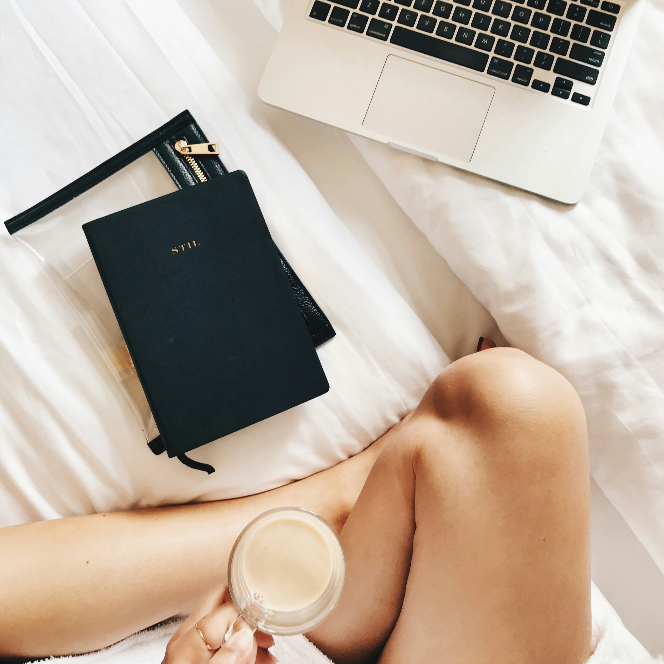 a top-down view of a woman holding a coffee cup, only her knee is visible, and a notebook and laptop sit on the bed in front of her. used in a blog post about wedding photography workflows.
