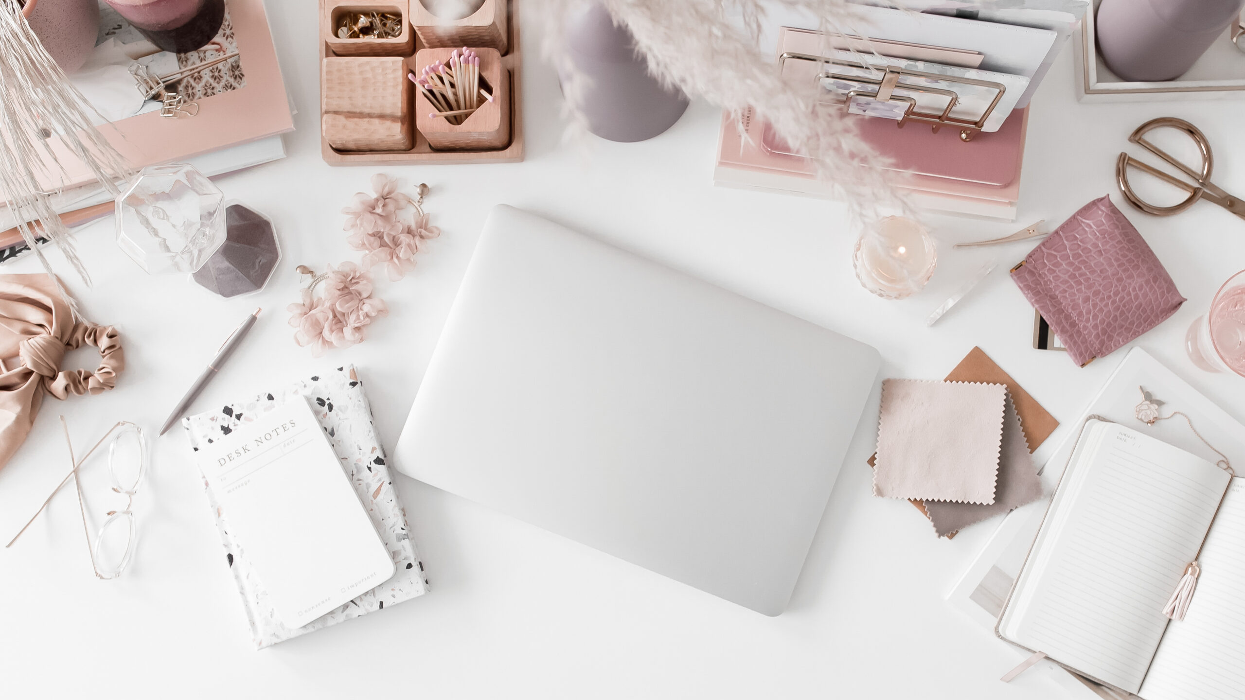 Flat lay of a white desk with a closed laptop, notebooks, glasses, and pink office supplies — stock image used for a blog on how to refresh wedding photography systems.